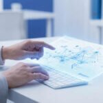 Copywriting IA : la stratégie pour des textes humains 1 Mains manipulant un clavier et un écran holographique lumineux affichant des visualisations de données complexes et un réseau neuronal. Contexte de bureau moderne.